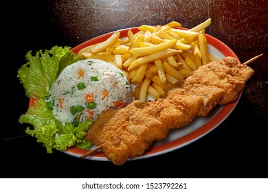 Breaded Fish Fillet Skewer With Chips, Greek Rice And Lettuce Leaves On A White Plate On A Wooden Table