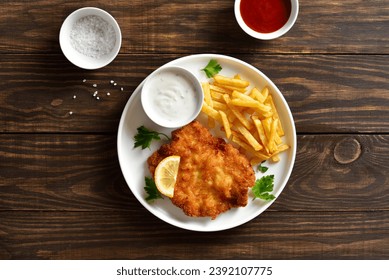 Breaded weiner schnitzel with potato fries and sauce on white plate over wooden background. Top view, flat lay