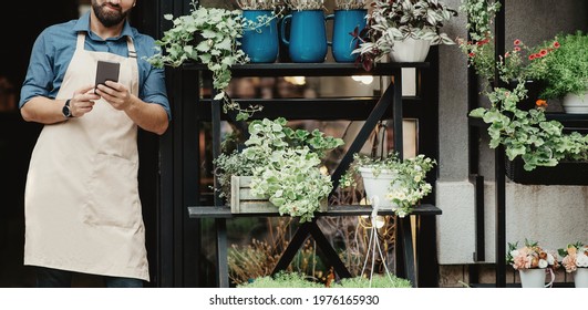 Break With Modern Device In Flower Studio. Young Smiling Bearded Caucasian Guy Seller In Apron Typing On Smartphone At Front Door Of Shop In Rustic Style With Different Plants, Panorama, Cropped