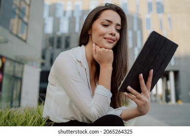 Break from office, smartly dressed girl sits on concrete outside in front of glass modern corporate building, ornamental plants around her, woman is holding a tablet, watching videos, series