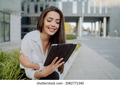 Break From Office, Smartly Dressed Girl Sits On Concrete Outside In Front Of Glass Modern Corporate Building, Ornamental Plants Around Her, Woman Is Holding A Tablet, Watching Videos, Series