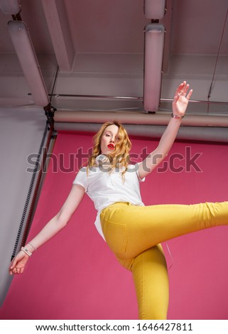 Break the rules. Attractive leggy blonde in yellow jeans poses in the studio on a pink background. Kick motion. Lower view.