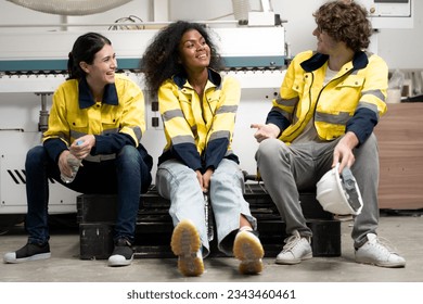 Break time. Group of diverse engineer people sitting together take a break and talking. Young multiracial woman and colleagues technician relax after job in factory. Manufacturing employee lifestyle.
