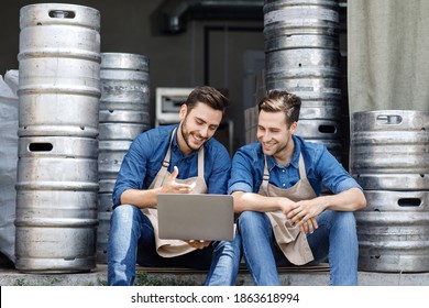 Break At Warehouse And Rest Of Employees. Happy Millennial Guys, Owners In Aprons Sit On Floor, Look At Data In Laptop And Talk About Business In Interior Of Warehouse With Metal Kegs, Free Space