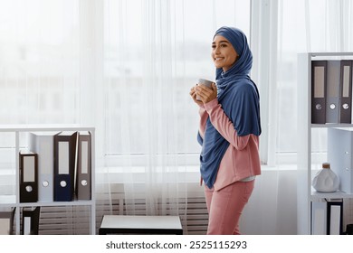 Break in work. Arabic female manager in hijab standing with coffee mug in office, relaxing against window at workplace, free space - Powered by Shutterstock