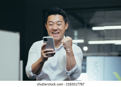 A break at work. A young Asian man holds a phone in his hands, plays games on the phone in the office at work, rejoices, shows with his hand yes,rejoices at the win, won a prize, rests, does not work.