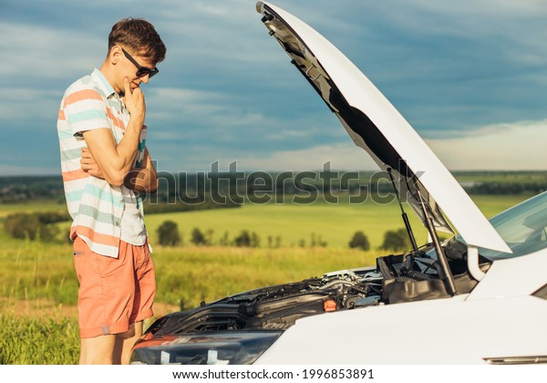 Breakdown of the car. Concentrated young man holding\
on to the hood of a car and looking inside while standing outdoors\
on a sunny day