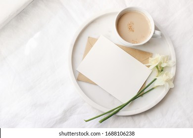 Breakfast in bed mockup scene near window. Cup of coffe, greeting card, narcissus, daffodil flowers on marble tray. White linen table cloth background. Spring, Easter concept. Flat lay, top view.