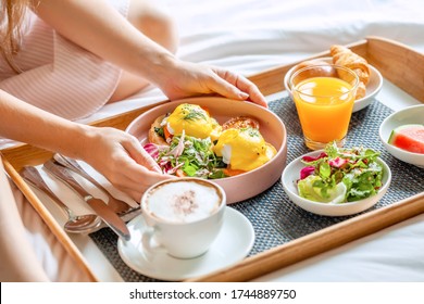 Breakfast in Bed Served with Cup of Coffee, Salad, Fresh Fruits and Eggs Benedict on Wooden Tray. Woman Hands Holding Plate with Fresh Food. Room Service in Hotel