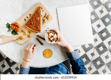 Breakfast In The Cafe. A Girl Is Sitting At A Table In A Cafe. Female Hands Hold Coffee With Cream. On The Table Is A Piece Of Cake And An Empty Blank, Mockup For Your Text. Top View