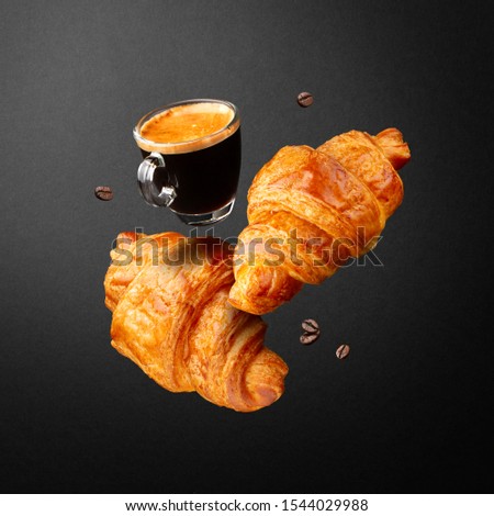 Breakfast or coffee house concept. Espresso in glass cup, croissants and beans coffee flying or falling in air on black background.