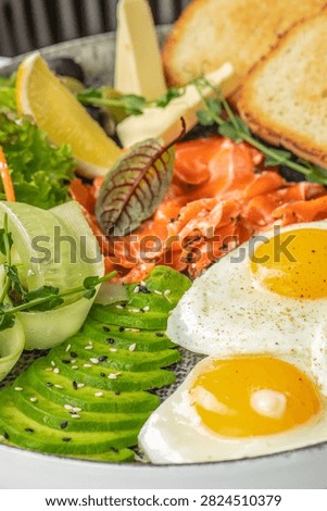 Breakfast dish includes two fried eggs, smoked salmon, avocado, fresh greens, and toasted bread with a lemon wedge on a decorative plate