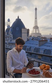 Breakfast, Eiffel Tower and tablet with man on balcony of hotel for holiday, travel or vacation. App, food and landmark with tourist on terrace of accommodation for sightseeing in Paris, France