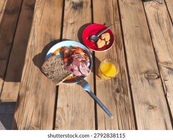 Breakfast featuring rye bread, sliced meats, tomatoes, cookies, and orange juice served on a wooden table in a bright outdoor environment