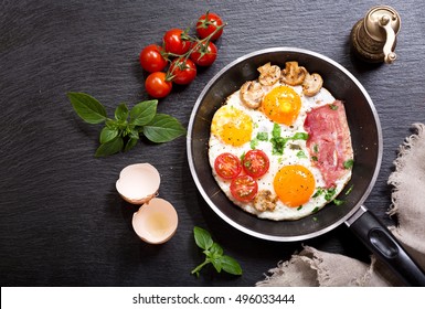 Breakfast With Fried Eggs In A Pan With Bacon And Vegetables On Dark Background, Top View