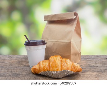 Breakfast To Go,coffee And Croissant With Paper Bag On Wooden Table Over Green Defocused Background