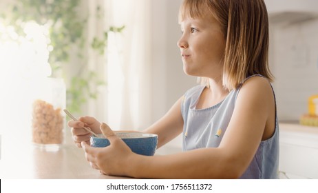 Breakfast In The Kitchen: Portrait Of Adorable Little Girl Eating Healthy Granola Cereal With Milk Out Of Bowl. Cute Little Girl Having Breakfast. In The Background Caring Mother Prepares Food