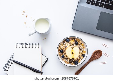 Breakfast In The Office With A Bowl Of Cereal, Fruit And Milk On A White Desk With A Laptop. Top View, Blank Spiral Notebook. 