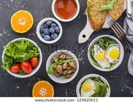Breakfast on a dark background: fried quail eggs, arugula, fresh bread, lettuce, tomatoes, oranges, nuts, tea, coffee fried mushrooms fresh blueberries Top view