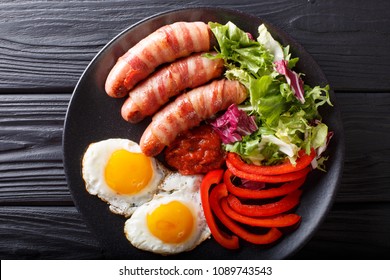 Breakfast Pigs In Blankets Fried Sausages Wrapped In Bacon, Eggs, Sauce And Fresh Salad Close-up On A Plate. Horizontal Top View From Above
