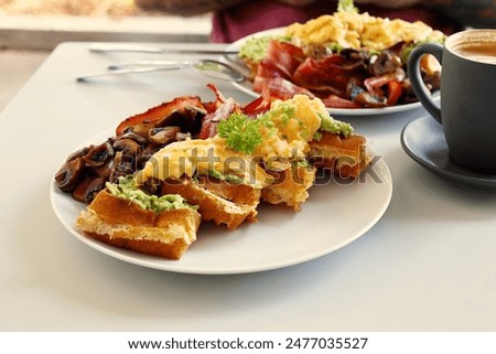 A breakfast platter with scrambled eggs, bacon, mushrooms, and waffles served with a black cup of coffee