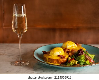 Breakfast In Restaurant  - Eggs Benedict With Hollandaise Cheese Sauce Served With Glass Of Champagne Wine In Restaurant Or Cafe Interior Background. Close Up, Selective Focus