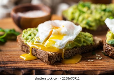 Breakfast Rye Bread Toast With Mushed Avocado And Poached Egg On Wooden Serving Board, Closeup View