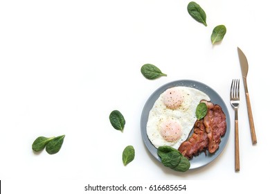 Breakfast served isolated on white top view. Eggs with bacon and fresh spinach on plate with fork and knife on table. Horizontal flatlay
