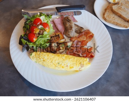 The breakfast set has rolled eggs, bacon, sausage, ham, roast potatoes and vegetable salad on a plate placed on the dining table. It looks very delicious and appetizing.         