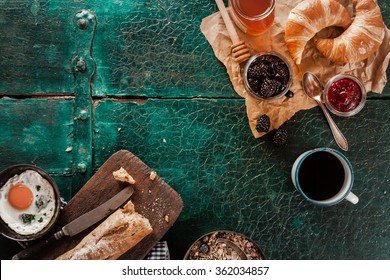 Breakfast Spread With Coffee, Bread, Fresh Croissants, Fried Egg, Honey And Preserves On An Old Rustic Green Wooden Table, Overhead View With Copyspace