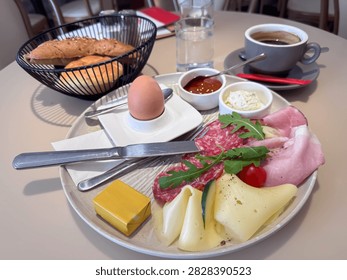 Breakfast spread includes a boiled egg, meats, cheeses, cherry tomato, and condiments, accompanied by bread rolls and coffee on a dining table