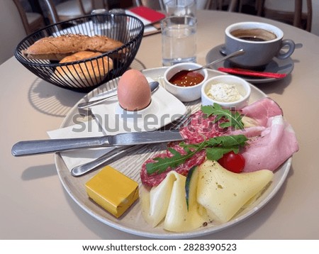 Breakfast spread includes a boiled egg, meats, cheeses, cherry tomato, and condiments, accompanied by bread rolls and coffee on a dining table