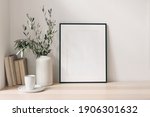 Breakfast still life. Cup of coffee, books and empty picture frame mockup on wooden desk, table. Vase with olive branches. Elegant working space, home office concept. Scandinavian interior design.
