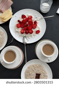 Breakfast table features cottage cheese with strawberries, oatmeal bowls, coffee cups, and a glass of milk on a dark surface