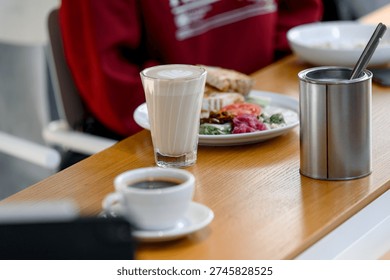 A breakfast table features a cup of coffee, a glass of latte, and a plate with various food items on a wooden surface