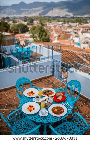 A breakfast table set on a rooftop terrace with a beautiful city and mountain view in the background. Plates of fresh fruit, coffee, and traditional Mexican dishes are served on a bright blue table.