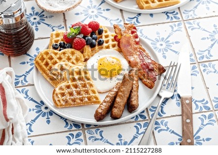 Breakfast table with waffles, fried egg, bacon and sausage served with fresh berries and maple syrup
