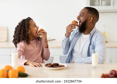 It's Breakfast Time. Portrait Of Cheerful Black Little Girl And Man Eating Sweet Muffin Cake Cookies And Looking At Each Other, Dad And Daughter Sitting At Table In Kitchen, Enjoying Meal With Milk