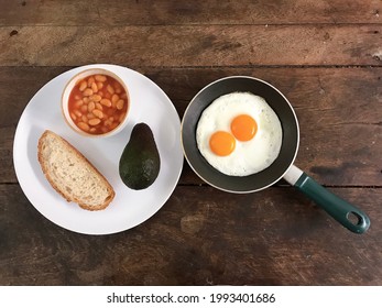 Breakfast Today Is Fried Eggs In A Pan And Piece Of Bread, One Avocado And A Bowl Of Baked Bean With Wood Table Background