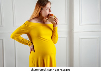 Breaking Diet Rules. Lady In Yellow Waiting For A Child And Eating A Fast Food Sandwich With An Appetite With White Wall On Background