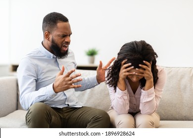 Breaking Up And Divorce Concept. Young married black couple having fight, guy yelling at crying lady, gesturing at home. Furious man shouting at his wife or girlfriend, sitting on sofa at living room