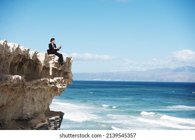 Breaking Free From The Rat-race. Young Businessman Sitting On The Edge Of A Cliff Overlooking The Ocean While Reading A Book.