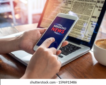 Breaking News Online On Smartphone. Finger Touching On Mobile Phone For Reading News Online On Smartphone With Laptop Computer And Coffee Cup On Wooden Table.