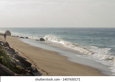 breaking wave on sandy beach, man walking leaving footprints into distance