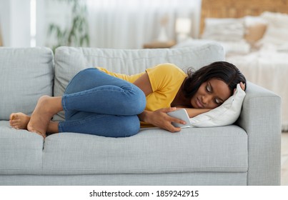 Breakup Depression Concept. Stressed Black Woman Lying On Sofa With Cellphone And Reading Bad Message At Home. Upset Young Lady With Mobile Phone Feeling Sad, Suffering From Loneliness On Web