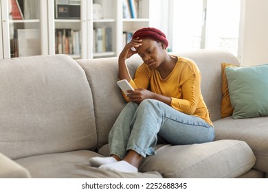 Breakup depression concept. Upset young black woman sitting on sofa with cellphone and reading bad message at home, female with mobile phone feeling sad, suffering from loneliness on web, copy space - Powered by Shutterstock