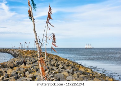 Breakwater Or Pier And Poles With Colored Rope And Plastic Waving In The Wind And A Ship With White Sails In The Wadden Sea At The Entrance To The Port Of Harlingen The Netherlands