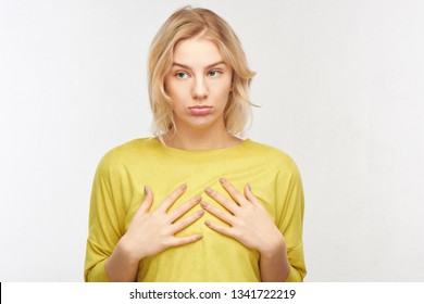 Breast augmentation concept. Upset young woman with a displeased facial expression touches her small size in the studio on a white background