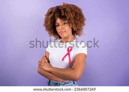 Breast Cancer Awareness. Black Young Woman With Pink Ribbon On T-Shirt Standing Over Purple Background In Studio, Crossing Hands Looking At Camera. Portrait Shot. Oncology Treatment