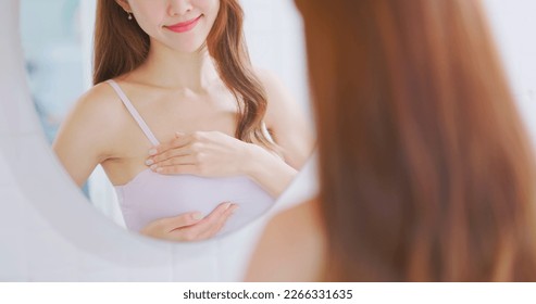breast cancer self check concept -  asian woman is touching her chest to prevent sick while looking in mirror at home - Shutterstock ID 2266331635